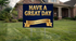 A rectangular yard sign with a dark navy blue background and large gold text reading “HAVE A GREAT DAY,” plus a gold banner reading “The Gold Standard Resolved,” displayed on a metal stand in a green lawn in front of a house.